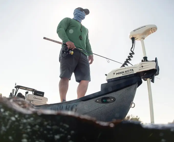 Fisherman on a boat equipped with Minn Kota trolling motor, dressed in a green shirt and shorts, face covered with a bandana, holding a fishing rod. The setting conveys an adventurous vibe.