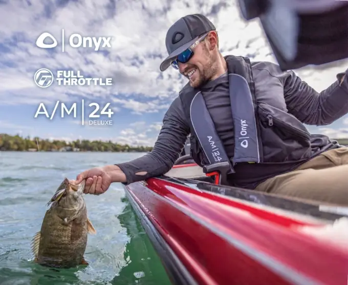 Man in a cap and sunglasses grins while pulling a large fish from a lake beside his red boat. He wears an Onyx life jacket under a clear sky.